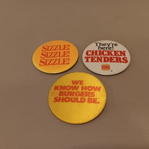May include: Three vintage Burger King pinback buttons. One is yellow with "SIZZLE!" repeated. Another is white with "They're here! CHICKEN TENDERS." The third is yellow with "WE KNOW HOW BURGERS SHOULD BE."