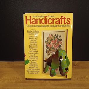 May include: A yellow book titled "The Complete Book of Handicrafts" with a step-by-step guide. The cover features a green and brown dog-shaped craft and a framed floral embroidery. The book lists various crafts like embroidery, crochet, and macrame.