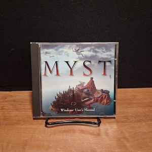 May include: A clear plastic CD jewel case containing the Myst Windows User's Manual. The cover art features a silhouette of a person falling against a cloudy sky above a detailed island landscape with various structures and the title text MYST.