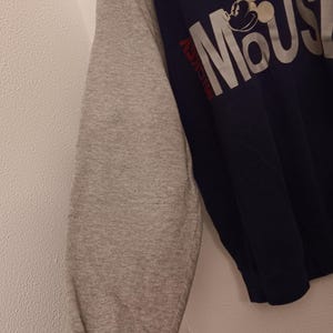 May include: Navy blue and gray raglan-sleeve sweatshirt featuring the word "MOUSE" in large white letters with a Mickey Mouse graphic. The sleeve is gray with a red and white striped cuff. The word "Mickey" is printed in red.