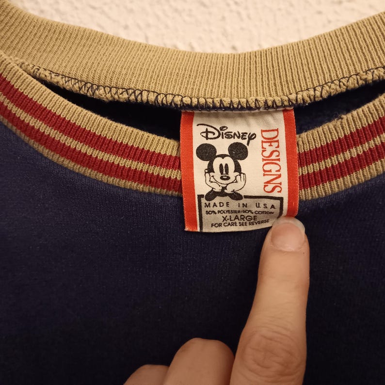 May include: A navy blue sweatshirt with a tan collar and red and white striped accents. The Disney Designs tag features Mickey Mouse and reads "MADE IN USA" and "X-LARGE".
