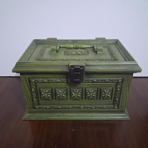 May include: A rectangular olive green storage box featuring a hinged lid with a central handle and a dark brown latch. The exterior is decorated with molded floral relief patterns and a wood grain texture on all visible sides.