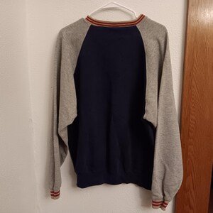 May include: A navy blue and gray raglan-sleeve sweatshirt hanging on a white hanger. The sweatshirt has a navy blue body, gray sleeves, and red and tan striped trim on the collar and cuffs. The garment is made of a soft material.