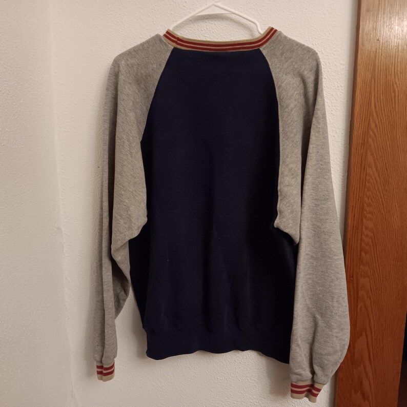 May include: A navy blue and gray raglan-sleeve sweatshirt hanging on a white hanger. The sweatshirt has a navy blue body, gray sleeves, and red and tan striped trim on the collar and cuffs. The garment is made of a soft material.