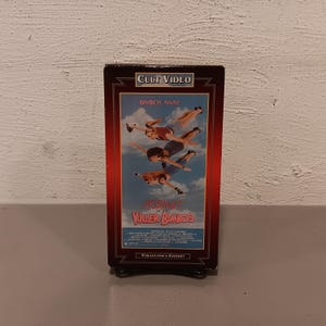 May include: A vintage VHS tape for "Assault of the Killer Bimbos" is displayed. The cover features a skydiving scene with the text "Bimbos Away!" and "Collector's Edition". The tape is in a black case with a red border.