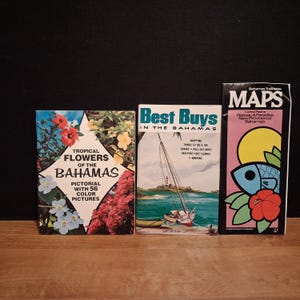 May include: Three travel guidebooks about the Bahamas. The left book features tropical flowers and text. The center book shows a sailboat on water with the title Best Buys in the Bahamas. The right book is titled Maps and features a graphic fish.
