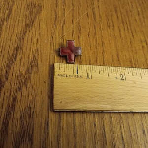 May include: A small, red cross-shaped pin with a textured design. The pin is next to a wooden ruler that reads "MADE IN U.S.A."