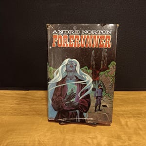 May include: A vintage paperback book titled "Forerunner" by Andre Norton. The cover art features a celestial figure with long white hair, holding a small image, against a backdrop of a starry sky and alien landscape. The book is displayed on a black stand.
