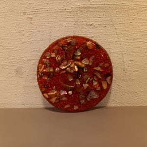 May include: A round, red resin coaster with embedded seashells and glitter. The coaster has a glossy finish and is approximately 4 inches in diameter. The seashells are various sizes and shapes, adding texture and visual interest to the coaster.