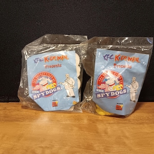 May include: Two sealed KFC Kids Meal toys. Each package features a blue background with the text "KFC Kids Meal Presents" and "The Secret Files of The Spydogs". The packages are in clear plastic.