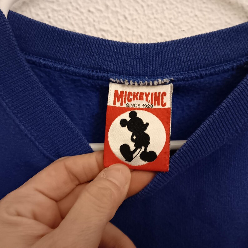 Pode incluir: Um close-up de um moletom azul com uma etiqueta que diz "MICKEY, INC. SINCE 1928". A etiqueta apresenta uma silhueta do Mickey Mouse em preto sobre um c&iacute;rculo branco, em um fundo vermelho. O moletom &eacute; azul s&oacute;lido e vibrante.