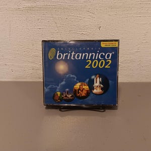 May include: A blue CD case with the text "Deluxe Edition CD" and "ENCYCLOPEDIA britannica 2002" on the front. The case features a variety of images, including a space shuttle, a globe, and a building.