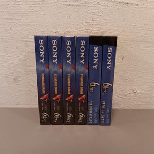 May include: A collection of six VHS video tapes. The tapes are dark blue with the brand name "SONY" in white. Some tapes are labeled "STANDARD GRADE" and others "PREMIUM GRADE". Each tape is labeled "6hrs".
