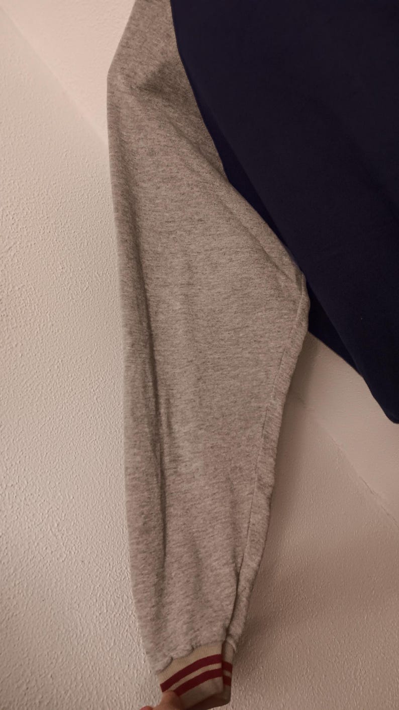 May include: A navy blue and gray long-sleeved shirt. The gray sleeve has a ribbed cuff with red and white stripes. The shirt is made of a soft material.
