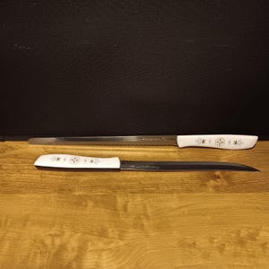 May include: Two knives with white handles and decorative starburst designs. One knife is a serrated bread knife, and the other is a carving knife. The blades are silver, and the handles are white with a starburst pattern.