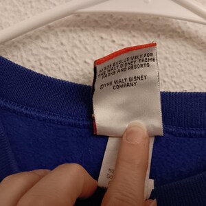 Pode incluir: Um close-up de uma pe&ccedil;a de roupa azul com uma etiqueta branca. A etiqueta diz "MADE EXCLUSIVELY FOR THE WALT DISNEY THEME PARKS AND RESORTS" e "THE WALT DISNEY COMPANY". A etiqueta &eacute; segurada por um dedo.