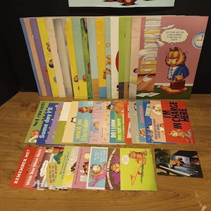 May include: A collection of Garfield comic books and cards. The books are stacked vertically, with various covers featuring Garfield. The cards are laid out in rows, each with a different Garfield illustration and text. The color palette includes orange, yellow, and blue.