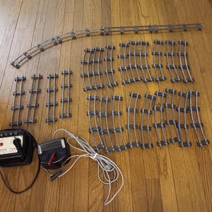 Può includere: Una collezione di binari per trenini modello vintage, tra cui sezioni dritte e curve, insieme a un trasformatore e un regolatore di alimentazione. I binari argentati sono disposti su una superficie di legno, suggerendo un classico set di trenini.