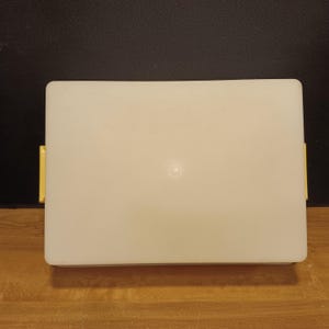 May include: A rectangular, off-white plastic container with rounded corners. It has two small, yellow handles on the sides. The container is set against a dark background.