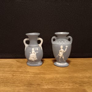 Pode incluir: Dois pequenos vasos de cerâmica azul-acinzentada com desenhos em relevo brancos. Um vaso apresenta uma mulher com um vestido esvoaçante, o outro um homem com uma espada. Ambos têm alças curvas e bases decorativas.