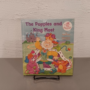 May include: A children's book titled "The Popples and King Most" featuring a cartoon illustration of a king with a crown, surrounded by colorful characters. The cover art includes a castle and the Popples logo.