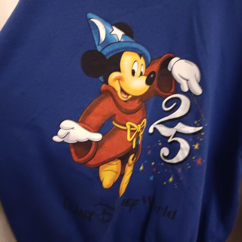 Pode incluir: Camisola azul com um desenho do Mickey Mouse como feiticeiro, usando um chap&eacute;u azul e uma t&uacute;nica vermelha. O n&uacute;mero "25" est&aacute; em branco, com "Celebrate Disney World" por baixo. Um design cl&aacute;ssico da Disney.