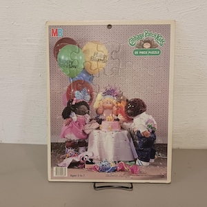 May include: A 25-piece jigsaw puzzle featuring the Cabbage Patch Kids celebrating a birthday. The puzzle shows three Cabbage Patch Kids dolls, two girls and one boy, with balloons and a birthday cake. The puzzle is designed for ages 3 to 7.