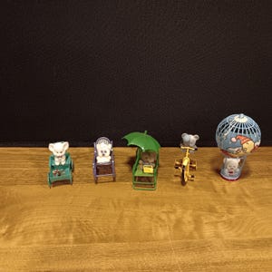 May include: A collection of miniature teddy bear figurines. One bear is in a green cart, another in a purple chair, and a third under a green umbrella. A fourth bear rides a yellow tricycle, and the last is in a blue and white hot air balloon.