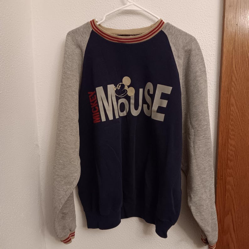 May include: A navy blue and gray raglan-sleeve sweatshirt featuring the text "Mickey Mouse" in white. The word "Mickey" is in red on the left side. The collar and cuffs have red and white stripes. A classic Disney-themed apparel item.