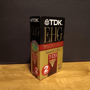 May include: A TDK EHG T-120 VHS tape two-pack. The box is black and gold with red accents. The text "EXTRA HIGH GRADE Sound & Color" is visible. The box has a globe graphic and the text "2 PACK".