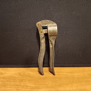 May include: A vintage, silver-toned garlic press with the brand name "SIMPLEX" stamped on the top. The press has curved handles and a hinged design for easy use. The tool is made of metal and shows signs of wear.