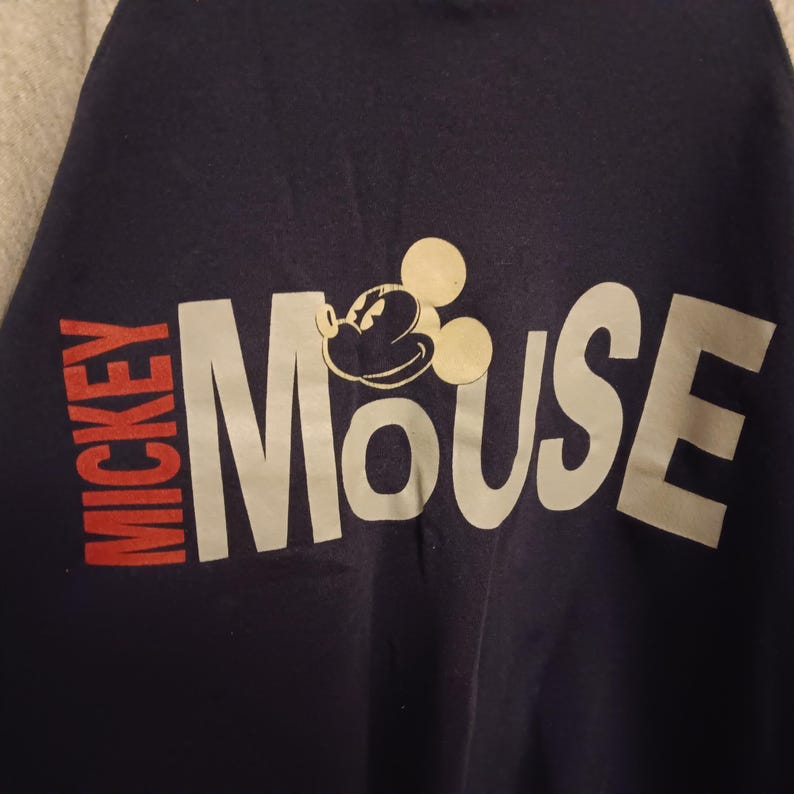 May include: Navy blue t-shirt featuring the text "Mickey Mouse" in white and red. The design includes a graphic of Mickey Mouse's face and ears. The shirt is a classic piece of Disney apparel.