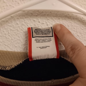 May include: Close-up of a tag with care instructions and the text "MADE EXCLUSIVELY FOR THE WALT DISNEY THEME PARKS AND RESORTS" and "THE WALT DISNEY COMPANY". The tag is red and white.