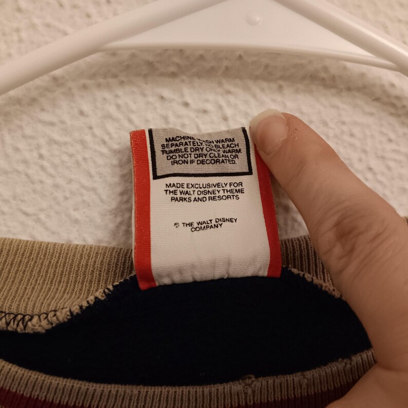 May include: Close-up of a tag with care instructions and the text "MADE EXCLUSIVELY FOR THE WALT DISNEY THEME PARKS AND RESORTS" and "THE WALT DISNEY COMPANY". The tag is red and white.