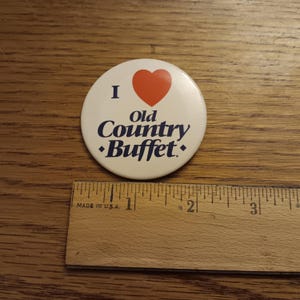 Pode incluir: Um botão redondo e branco com o texto "I ❤️ Old Country Buffet" em azul e um coração vermelho. O botão está sobre uma superfície de madeira ao lado de uma régua de madeira.