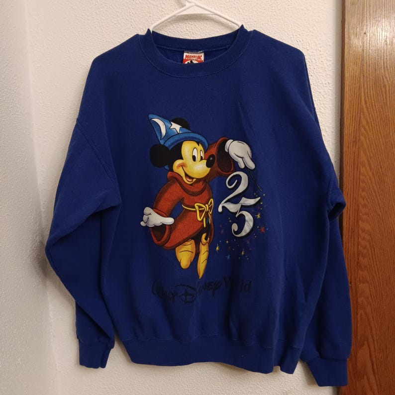 Pode incluir: Um moletom azul royal com um gr&aacute;fico do Mickey Mouse em um traje de mago, com o n&uacute;mero 25 e as palavras "Our Disney World". O moletom tem gola redonda e mangas compridas.