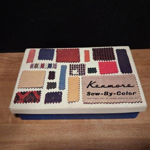 Könnte beinhalten: Eine rechteckige weiße Schachtel mit blauem Boden, die verschiedene Stoffmuster in roten, blauen, schwarzen und beigen Mustern zeigt. Der Deckel trägt den Text Kenmore Sew-By-Color und Copyright 1960 by Sears Roebuck and Co. in schwarzer Schrift.