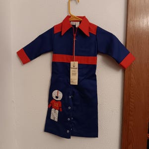 Könnte beinhalten: Ein blauer und roter Vintage-Overall für ein Kind, an einem gelben Kleiderbügel aufgehängt. Der Overall hat einen roten Kragen, Manschetten und einen horizontalen Streifen über der Brust. Eine Cartoonfigur mit der Nummer 12 befindet sich unten links.