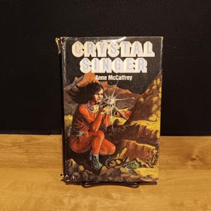 May include: A vintage paperback book titled "Crystal Singer" by Anne McCaffrey. The cover art features a woman in a red jumpsuit holding a glowing object, set against a rocky, desert-like landscape. The title is in large, white, block letters.