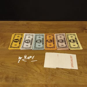 May include: A collection of play money in various denominations, including $1, $5, $10, $20, $50, and $100 bills. Also includes white game pieces and a stack of cards with the word "SPECIALIST" printed on them. The items are arranged on a wooden surface.