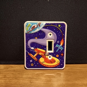 May include: A decorative light switch cover featuring a retro space-themed design. The cover has a purple background with a planet, stars, and a spaceship. The switch opening is rectangular, and the cover has a white border.