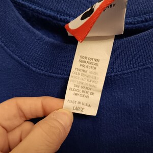 Pode incluir: Close-up de um moletom azul royal com uma etiqueta. A etiqueta diz "50% Algod&atilde;o, 50% Fortrel Poli&eacute;ster, Lavar &agrave; m&aacute;quina separadamente com &aacute;gua fria, Made in U.S.A., Large". A etiqueta tamb&eacute;m tem um design vermelho e branco.