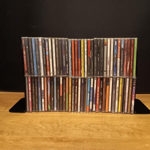May include: A collection of numerous music CDs arranged in two horizontal rows within a black metal rack. The spines of the jewel cases display various colors, text, and graphic designs, showcasing a diverse assortment of musical artists and albums.