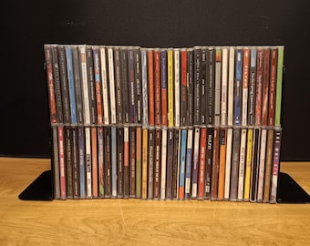 May include: A collection of numerous music CDs arranged in two horizontal rows within a black metal rack. The spines of the jewel cases display various colors, text, and graphic designs, showcasing a diverse assortment of musical artists and albums.