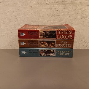 May include: Three stacked paperback books with titles "Fortress Draconis", "When Dragons Rage", and "The Grand Crusade". The books are in shades of red, brown, and blue, with author Michael A. Stackpole's name visible.