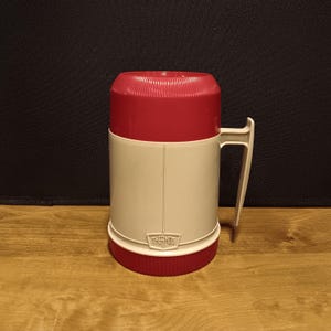 May include: A vintage Thermos brand insulated mug. The mug is primarily off-white with a red lid and base. It has a white handle and the Thermos logo is visible on the front. The mug is designed to keep beverages hot or cold.