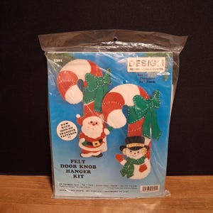 May include: A Design Works Crafts felt door knob hanger kit in a clear plastic bag. The kit features two candy cane designs, one with a Santa Claus and one with a snowman, each with green bows. Text includes Set of Two Finishes 11 inches Each.
