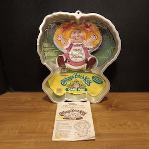 May include: A silver-colored aluminum Cabbage Patch Kids cake pan with a cardboard insert featuring a decorated doll cake and the text Happy Birthday Lauren. Below the pan is a folded paper instruction booklet with the Cabbage Patch Kids logo.
