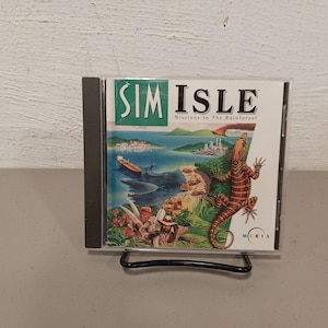 May include: A CD case for the computer game SimIsle: Missions in the Rainforest. The case features a colorful illustration of a tropical island with a lizard, a boat, and two people.
