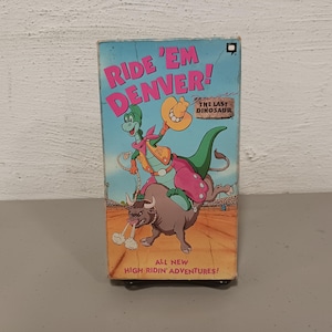 May include: A VHS tape featuring the cartoon character 'Ride 'Em Denver! The Last Dinosaur'. The cover shows a green dinosaur wearing a cowboy hat and riding a brown bull. The text 'All New High Ridin' Adventures!' is at the bottom of the cover.